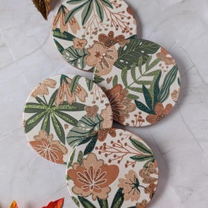 May include: A set of four circular coasters featuring a botanical pattern with cream, sage green, and terracotta orange floral and leaf motifs. The design includes shimmering glitter accents on the petals and foliage, creating a textured appearance.