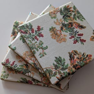 Könnte beinhalten: Stapel von vier quadratischen Untersetzern mit floralem Design. Die Untersetzer haben einen weißen Hintergrund mit einem Muster aus grünen, orangefarbenen und roten Blättern und Beeren. Die Ränder sind mit einem goldenen Rand versehen. Jede Seite misst ca. 10 cm.