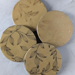 May include: Four circular coasters with a tan base colour, each featuring a delicate line-art botanical pattern of leaves and stems. Two coasters display the design in black ink, while the other two feature the same pattern in metallic gold.