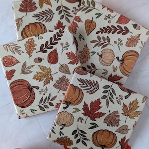 Puede incluir: Cuatro posavasos cuadrados de cerámica con un patrón repetitivo de calabazas naranjas, bellotas marrones y varias hojas de otoño en tonos rojos, dorados y marrones. El diseño incluye sutiles detalles de purpurina en las hojas.