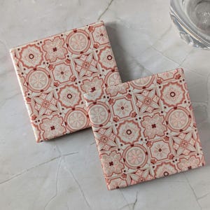 Könnte beinhalten: Zwei quadratische Untersetzer mit einem sich wiederholenden geometrischen Muster in Altrosa, zartem Rosa und Weiß. Jeder Untersetzer zeigt ein Gitter aus komplizierten Blumen- und sternartigen Motiven mit einer glänzenden Oberfläche.