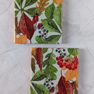 Könnte beinhalten: Zwei quadratische Untersetzer mit einem botanischen Druck aus Herbstblättern, roten Beeren und dunkelblauen Beeren auf hellem Grund. Die Oberfläche der Untersetzer hat ein glänzendes Finish mit dezenten Glitzerakzenten im Blattdesign.