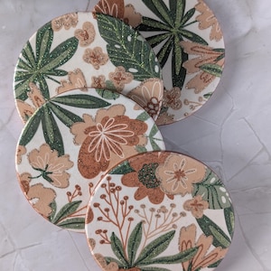 May include: Three circular coasters featuring a botanical pattern with cream backgrounds, dark green leaves, and tan flowers. The design includes shimmering glitter accents on the foliage and petals, with each coaster having a smooth, rounded edge.