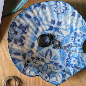 Op de afbeelding: Een decoratieve, schelpvormige sieradenschaal met een blauw-wit bloemmotief. De schaal bevat een zilveren ring en een hartvormige hangerketting. De achtergrond toont een houten oppervlak en een wenskaart.