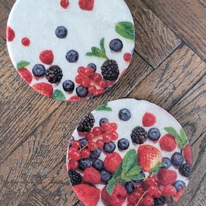 Könnte beinhalten: Zwei kreisförmige Untersetzer mit weißem Hintergrund, verziert mit einem lebendigen Muster aus frischen Beeren wie Erdbeeren, Blaubeeren, Brombeeren und Johannisbeeren, akzentuiert durch kleine, verstreute grüne Minzblätter.