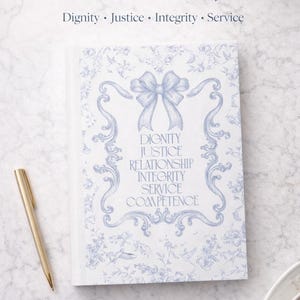 Op de afbeelding: Een dagboek getiteld "Social Work Core Values Journal" met een lichtblauw bloemmotief en de woorden "Dignity, Justice, Integrity, Service". Een gouden pen ligt naast het dagboek op een marmeren oppervlak. De kaft van het dagboek heeft een strik en de woorden "Dignity, Justice, Relationship, Integrity, Service, Competence."