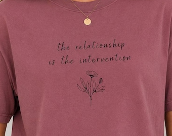 May include: A mauve short-sleeved t-shirt featuring the black printed text "the relationship is the intervention" centered on the chest, positioned above a simple black line-art illustration of a single flower with leaves and a bud.