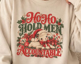May include: A beige crewneck sweatshirt featuring a graphic print of a lamb wearing a Santa hat and a red bow. The text reads Ho Ho Hold Men Accountable in red and green lettering, surrounded by holly, berries, and candy cane illustrations.