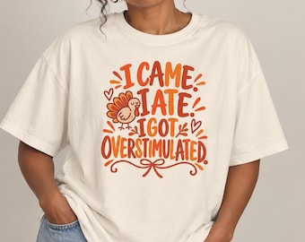 Peut inclure: Un t-shirt à manches courtes blanc cassé avec une impression graphique dans des tons orange et rouge. Le texte indique I CAME I ATE I GOT OVERSTIMULATED, accompagné d'une petite illustration de dinde et de motifs décoratifs et cœurs.