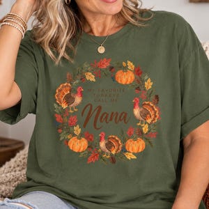 May include: An olive green short-sleeve t-shirt featuring a circular autumn wreath graphic with pumpkins, fall leaves, and three turkeys. The center text reads, My Favorite Turkeys Call Me Nana, in a combination of block and script lettering.