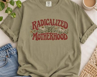 May include: An olive green short-sleeve t-shirt featuring the red text "RADICALIZED BY MOTHERHOOD" arched over a graphic of white daisies and green leaves. The shirt has a crew neckline and a tag at the inner collar.
