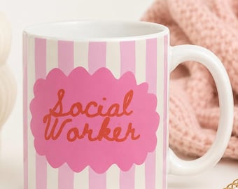 Puede incluir: Una taza de cerámica blanca con rayas verticales de color rosa claro y una forma ovalada festoneada de color rosa en el centro. El texto Social Worker está impreso en una fuente cursiva naranja-roja en el centro del óvalo rosa frontal.