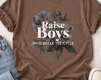 May include: A brown short-sleeve t-shirt featuring a black and white floral graphic with the white text "Raise Boys WHO BREAK THE CYCLE" printed in the center. The shirt has a crew neckline and a Comfort Colors brand label at the inner neck.