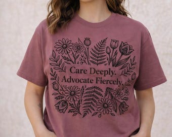 Puede incluir: Una camiseta de manga corta color malva con un estampado gráfico negro de varias flores y helechos que rodea el texto "Care Deeply. Advocate Fiercely." La camiseta tiene cuello redondo y un corte estándar, combinada con pantalones vaqueros azules.