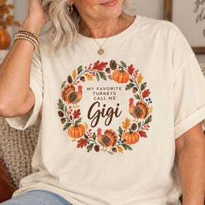 May include: A cream-colored short-sleeve t-shirt featuring a circular autumn-themed wreath graphic. The design includes pumpkins, fall leaves, acorns, and three turkeys surrounding the text: My Favorite Turkeys Call Me Gigi in brown lettering.