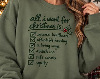 May include: An olive green crewneck sweatshirt featuring dark green text that reads: all i want for christmas is: followed by a checklist with checkmarks next to universal healthcare, affordable housing, a living wage, abolish ice, safe schools, and equity.