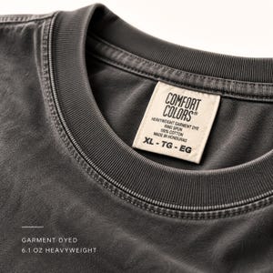 Puede incluir: Una camiseta gris carb&oacute;n con cuello redondo acanalado y una etiqueta interior color crema que dice COMFORT COLORS, HEAVYWEIGHT GARMENT DYE, RING SPUN, 100% COTTON, MADE IN HONDURAS, XL-TG-EG. El texto inferior dice GARMENT DYED 6.1 OZ HEAVYWEIGHT.