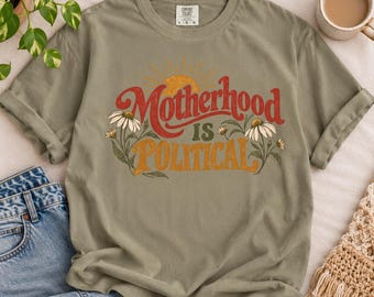 May include: An olive green short-sleeve t-shirt featuring the text "Motherhood IS POLITICAL" in red and orange script and block lettering. The design includes a yellow sun graphic and white daisy illustrations with green stems and leaves.