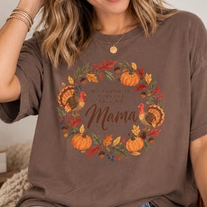 May include: A brown short-sleeve t-shirt featuring a circular autumn-themed wreath graphic. The design includes two turkeys, orange pumpkins, acorns, and fall leaves. The text in the center reads, "MY FAVORITE TURKEYS CALL ME Mama" in brown.