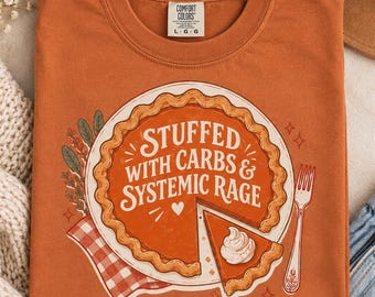 May include: A burnt orange crew neck t-shirt featuring a graphic of a pumpkin pie with a slice removed. The text reads STUFFED WITH CARBS & SYSTEMIC RAGE. The design includes a fork, a red and white checkered napkin, and sprigs of herbs.