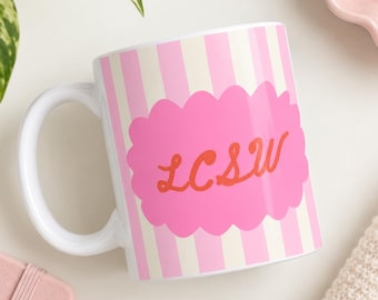 May include: A white ceramic mug featuring vertical light pink and cream stripes. Centered on the mug is a bright pink scalloped oval shape containing the orange cursive text LCSW. The mug has a curved handle on the left side.
