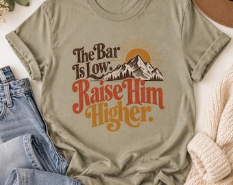May include: A sage green short-sleeve t-shirt featuring a graphic of a mountain range, a sun, and the text "The Bar Is Low. Raise Him Higher." The mountain and sun design is rendered in brown and orange tones, with the text in brown and orange.