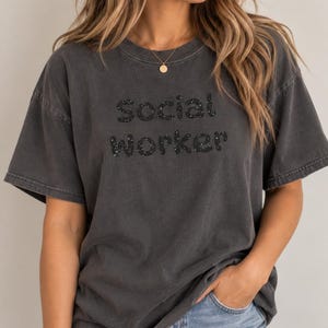 Könnte beinhalten: Ein anthrazitfarbenes Kurzarm-T-Shirt mit dem Schriftzug Social Worker in schwarzer Glitzerschrift auf der Brust. Das Kleidungsstück hat einen Rundhalsausschnitt und eine lockere Passform aus verwaschenem Stoff mit einer Vintage-Optik.