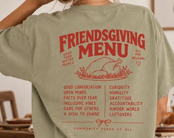 May include: A sage green t-shirt featuring red text and graphics on the back. The design includes the words "FRIENDSGIVING MENU," a turkey illustration, and a list of values like "GOOD CONVERSATION," "GRATITUDE," and "COMMUNITY FEEDS US ALL."