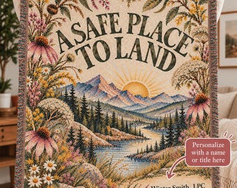 May include: A woven throw blanket featuring a landscape of mountains, a river, and a sunrise, framed by wildflowers. The text reads A SAFE PLACE TO LAND and Winter Smith, LPC. The blanket has a fringed edge and a multi-colored woven texture.