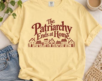 May include: A pale yellow short-sleeve t-shirt featuring the dark red text 'The Patriarchy Ends at Home' above a graphic illustration of a row of various houses. The shirt has a crew neckline and a visible brand label at the inner collar.