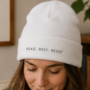 May include: A white beanie with the words "READ. REST. RESIST" printed in black. The beanie is made of a soft, knitted material and has a folded brim. The beanie is a comfortable and stylish accessory.