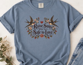 May include: A dusty blue short-sleeve t-shirt featuring a graphic of two swallows, floral vines, and a small red heart. The text reads, "Raise Sons Who Are Safe to Love." The shirt has a crew neckline and a Comfort Colors brand label at the neck.