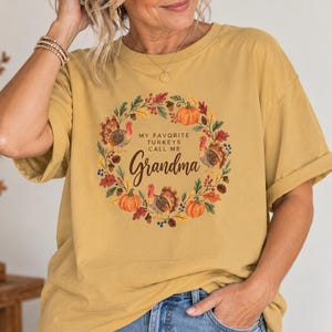 May include: A mustard yellow short-sleeve t-shirt featuring a circular wreath graphic made of autumn leaves, pumpkins, and turkeys. The center text reads, "MY FAVORITE TURKEYS CALL ME GRANDMA" in a dark brown font with a script style for the word Grandma.