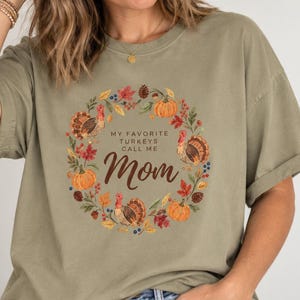 May include: An olive green short-sleeve t-shirt featuring a central graphic of a wreath made of autumn leaves, pumpkins, pinecones, and turkeys. The text in the center reads, "MY FAVORITE TURKEYS CALL ME Mom" in a mix of block and script fonts.