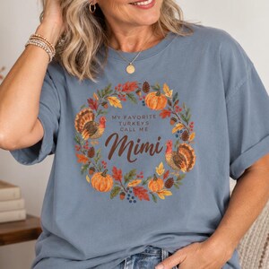 May include: A dusty blue short-sleeve t-shirt featuring a circular autumn-themed graphic. The design includes pumpkins, fall leaves, pinecones, and two turkeys surrounding the brown text that reads: MY FAVORITE TURKEYS CALL ME Mimi.