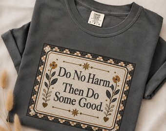 May include: A charcoal gray crew neck t-shirt featuring a rectangular graphic with a mosaic-style border. The text reads "Do No Harm. Then Do Some Good." and is flanked by two floral stems with gold-toned flowers and heart-shaped leaves.