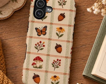 May include: A cream-colored phone case featuring a grid pattern with embroidered motifs including orange butterflies, brown acorns, red berries, yellow flowers, and red-and-white mushrooms. The case has a dual-lens camera cutout.