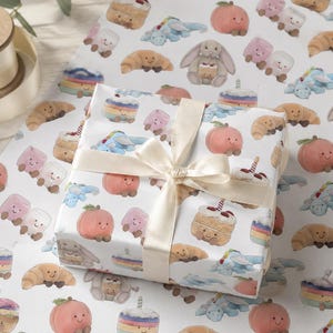 May include: White gift wrap with a repeating pattern of smiling baked goods and creatures, including cakes, croissants, peaches, marshmallows, and a bunny holding a cupcake. The wrapped gift is tied with a cream-colored ribbon.