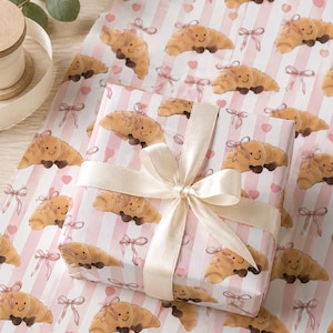 May include: Gift wrapping paper featuring a repeating pattern of smiling croissant illustrations with pink bows and hearts on a striped background. A wrapped gift with a cream-colored ribbon is in the center.