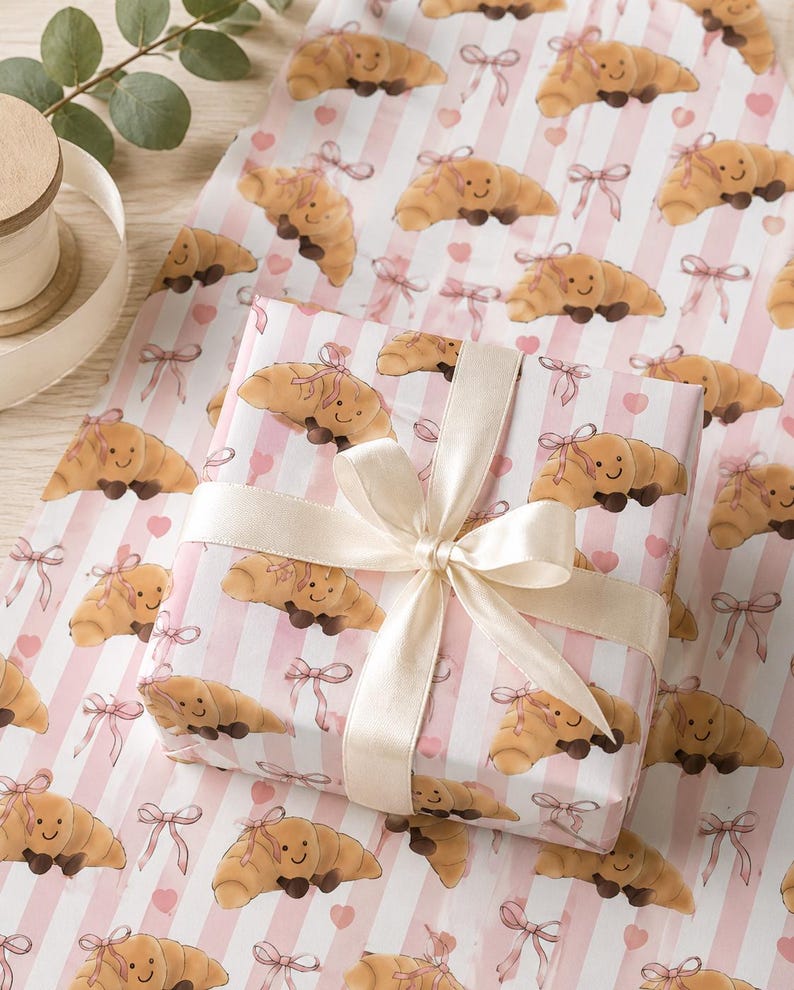 May include: Gift wrapping paper featuring a repeating pattern of smiling croissant illustrations with pink bows and hearts on a striped background. A wrapped gift with a cream-colored ribbon is in the center.