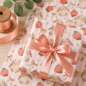 May include: A wrapped gift with a peach-colored satin ribbon and bow. The wrapping paper features a pattern of bunnies, peaches, and bows on a white and pink striped background. A spool of peach ribbon is in the upper left corner.