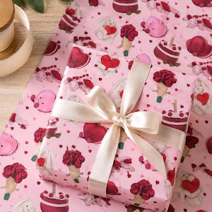 May include: A wrapped gift with a cream-colored ribbon bow sits on pink wrapping paper. The paper features a repeating pattern of cartoon hearts, bunnies, cakes, and ice cream cones. The gift wrap is perfect for Valentine's Day or a romantic occasion.
