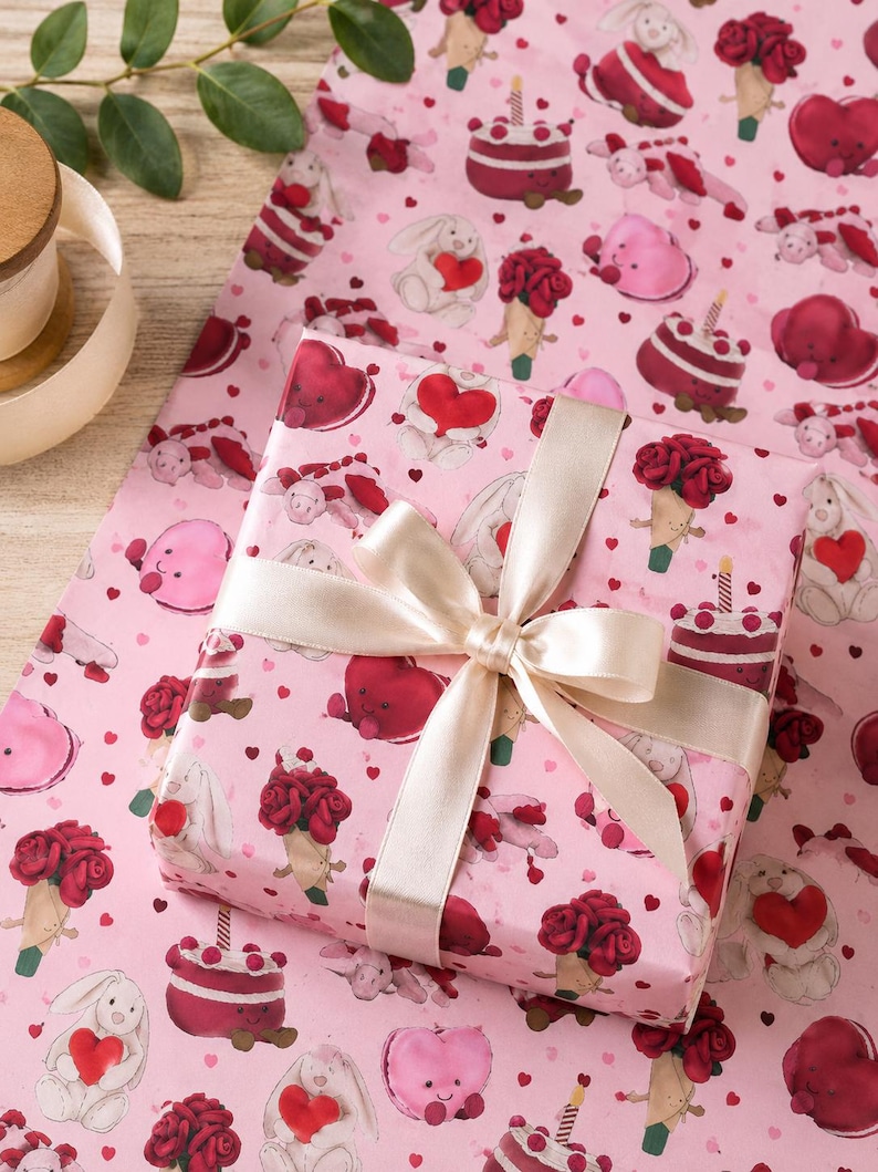 May include: A wrapped gift with a cream-colored ribbon bow sits on pink wrapping paper. The paper features a repeating pattern of cartoon hearts, bunnies, cakes, and ice cream cones. The gift wrap is perfect for Valentine's Day or a romantic occasion.