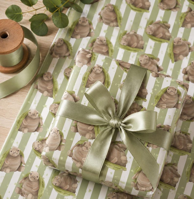 May include: Gift-wrapped present with a sage green satin bow, placed on matching wrapping paper. The paper features a repeating pattern of cartoon turtles on a white and green striped background. A wooden spool of ribbon and greenery are in the background.