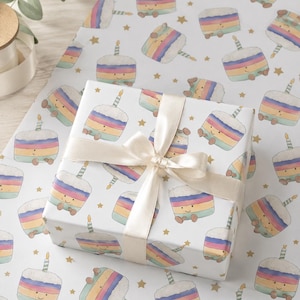 May include: A wrapped gift box with a cream-colored ribbon sits on a sheet of birthday-themed wrapping paper. The paper features a repeating pattern of smiling cartoon cakes with rainbow layers, candles, and gold stars.