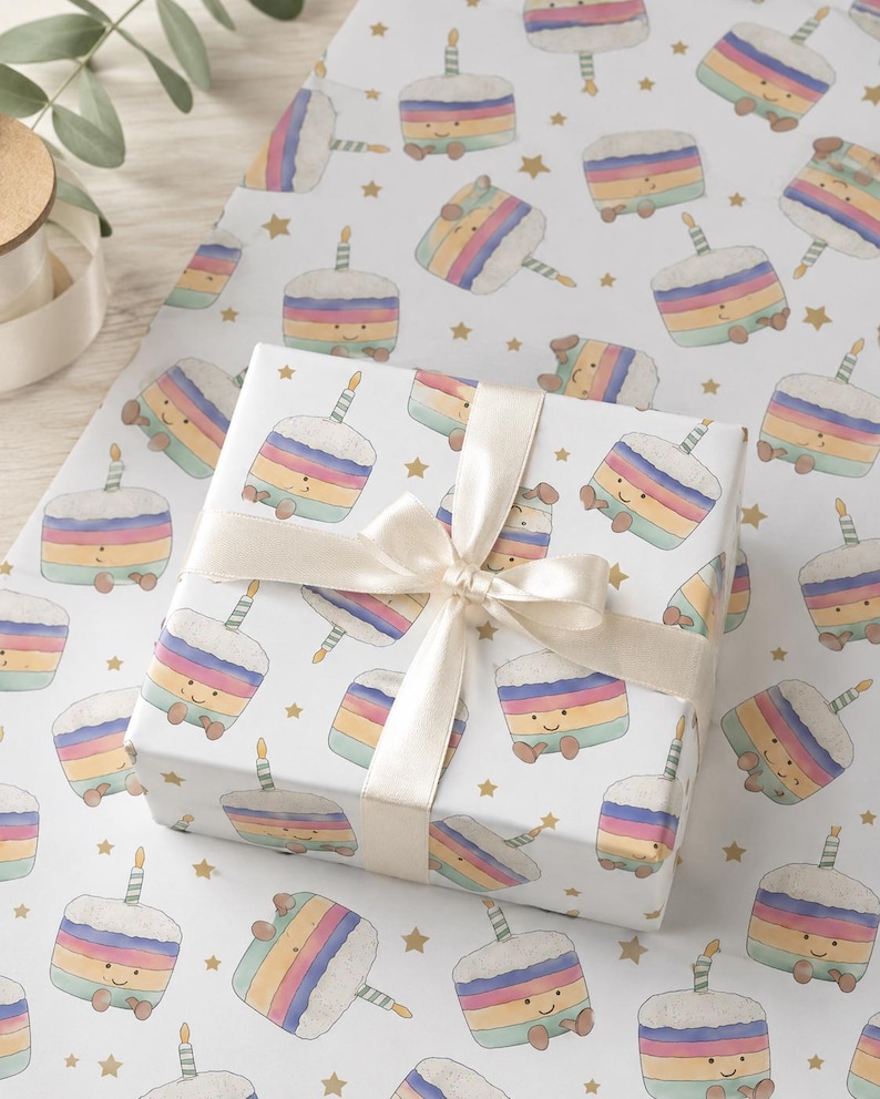 May include: A wrapped gift box with a cream-colored ribbon sits on a sheet of birthday-themed wrapping paper. The paper features a repeating pattern of smiling cartoon cakes with rainbow layers, candles, and gold stars.