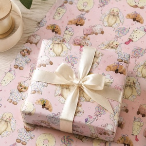 May include: A wrapped gift with a cream-colored satin ribbon bow sits on pink wrapping paper. The paper is patterned with illustrations of teddy bears, bunnies, donuts, and lollipops. The gift wrap is a light pink color.