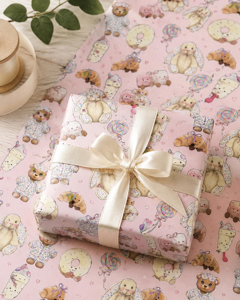 May include: A wrapped gift with a cream-colored satin ribbon bow sits on pink wrapping paper. The paper is patterned with illustrations of teddy bears, bunnies, donuts, and lollipops. The gift wrap is a light pink color.