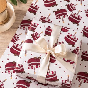 May include: A wrapped gift with a cream-colored ribbon sits on wrapping paper. The paper features a repeating pattern of cartoon cakes with candles and stars on a white background. The ribbon is tied in a bow.