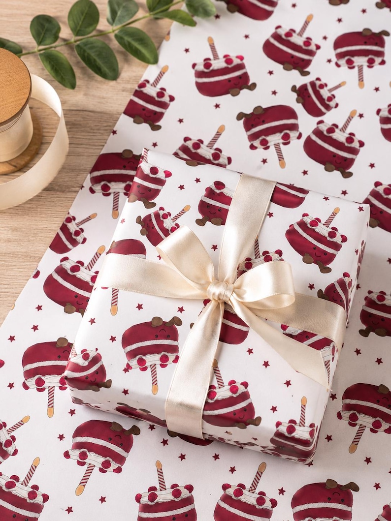 May include: A wrapped gift with a cream-colored ribbon sits on wrapping paper. The paper features a repeating pattern of cartoon cakes with candles and stars on a white background. The ribbon is tied in a bow.